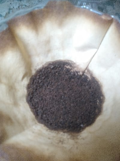 The image displays a close-up, overhead view of a used, light brown paper coffee filter containing dark brown, granular coffee grounds. The filter exhibits a crinkled texture and signs of moisture saturation, indicated by darker brown staining and dampness, particularly around its upper perimeter and beneath the central mass of grounds. The coffee grounds form an irregular, roughly circular mound in the center of the filter, appearing wet and compacted. The filter is positioned within what appears to be a transparent or translucent vessel, possibly glass, evidenced by faint greenish-blue reflections and subtle visual distortions visible along the filter's edges. No individuals or specific identifying objects are present. The scene is depicted as occurring within Ciudad de San José, Costa Rica, though no specific geographical markers are discernible within the image itself. The immediate environment suggests an indoor setting.