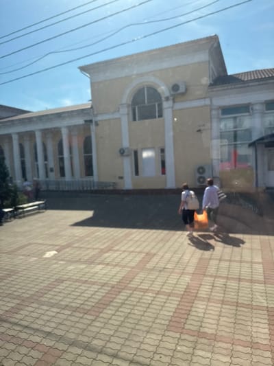 The image depicts the exterior of the Dagomys Railway Station, located in the Kholodnyi Rodnik microdistrict of Sochi, Krasnodar Krai, Russia, under a clear, bright blue sky with overhead utility lines. The central subject is a light yellow building with a classical architectural style, featuring a prominent facade with white trim, several tall arched windows, and a section supported by multiple white columns. Several external air conditioning units are mounted on the building's walls. The roof is reddish-brown and tiled. The ground in the foreground is paved with grey interlocking pavers arranged in a wavy pattern, interspersed with reddish-brown rectangular sections forming linear patterns. Strong sunlight from the left casts distinct shadows of individuals on the pavement. Two adult figures are walking away from the viewer towards the right side of the building. The person on the left wears a light top and dark bottom, carrying a light-colored backpack. The person on the right wears a light purple long-sleeved top and dark bottom, carrying a bright orange bag. In the far left background, partially obscured by foliage, dark benches are visible, with indistinct figures seated or standing nearby.