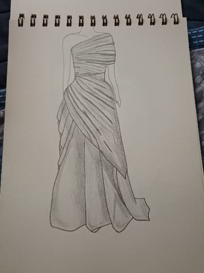 This image, captured in 서대문구, South Korea, depicts a graphite pencil drawing of a formal gown presented as a fashion illustration. The drawing occupies the majority of a white sheet of paper, which is part of a spiral-bound sketchbook with black metal coils visible along its top edge. The gown is modeled on a minimalist, headless, and armless figure.
The dress design features a one-shoulder neckline with intricate draping across the bodice, creating horizontal folds and a cinched waist. The full, floor-length skirt is characterized by multiple layers and elegant drapes, with an outer layer sweeping diagonally from the waist to the hem, forming a slight train. Shading with pencil defines the fabric's volume, texture, and the fall of the drapes, indicating depth and contour. The drawing technique is consistent throughout, employing varying pressure to achieve different shades of gray. The paper rests on an indistinct surface, with a dark, patterned fabric visible in the blurred periphery, particularly on the left and right sides of the page.