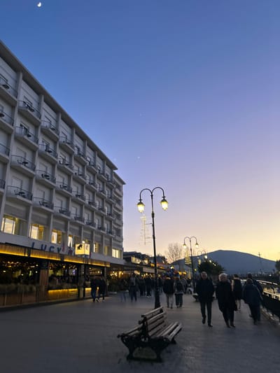 The image captures an outdoor scene at dusk along a promenade in Chalkida, Greece. The sky displays a gradient, transitioning from deep blue at the top, where a distinct crescent moon is visible, to a warm orange and yellow hue near the horizon.
On the left, a multi-story building, possibly a hotel or residential complex, features numerous balconies with railings and illuminated windows. The ground floor houses a commercial establishment with bright interior lights, and the text "LUCY" is visible on its signage.
To the right, a wide promenade is populated by several people strolling leisurely, either alone or in small groups. An ornate, double-headed streetlight with warm yellow lights illuminates the area, and festive string lights are draped around it and along the commercial facade. A dark park bench with metal armrests sits unoccupied further down the promenade. In the distance, dark silhouettes of mountains rise against the twilight sky. The scene suggests a calm and pleasant early evening atmosphere.