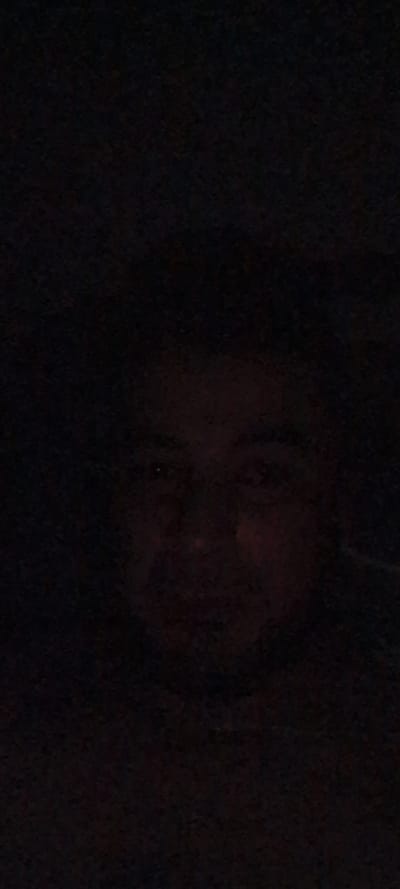 The media file is a dark, low-light photograph featuring what appears to be a person's face, possibly a child's, as the central subject. The face is mostly obscured by shadow, with only faint outlines and hints of features visible. A reddish glow emanates from around the face, suggesting an artificial light source or a reflection. The background is entirely dark and indistinct, providing no context for the setting, time of day, or weather conditions. There is no visible text in the image. The overall impression is one of mystery and obscurity, with the subject's identity and the surrounding environment remaining largely unknown. The provided location context of "Металлург Снт, Russia" does not offer any visual cues within the image itself.