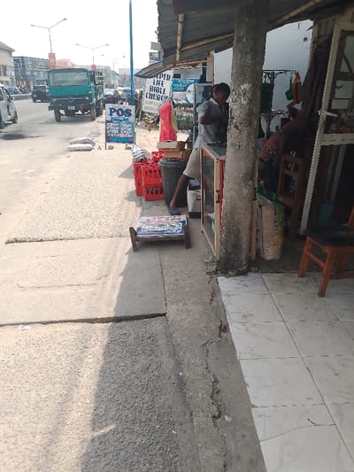 This eye-level outdoor shot depicts a bustling street scene in Mgbuoshimini, Nigeria, under bright daylight with strong shadows, indicating a sunny day. On the left, a paved road shows active traffic, including a prominent green Mercedes-Benz truck, a white van, and other vehicles. Street lights are visible in the background.

The right side of the image features a lively commercial sidewalk adjacent to a building with an extended awning. A man in a grey shirt and light shorts is partially visible under the awning, seemingly attending to a small glass display case or counter. He appears to be working at a street-side shop, with his back partially turned. Further inside the shop, another person is faintly visible near what might be a mannequin or clothing.

The sidewalk is cluttered with various objects associated with street commerce. A prominent blue "POS" (Point of Sale) sign advertises services such as "Cash Deposit," "Cash Withdraw," "Transfer," "Bill Payment," "Airtime," and payments for "Gotv," "Startimes," "Dstv," "Waec," "Jamb," "NECO," "Insurance," and "Betting." Behind it, a white sign partially reveals "KEPER LIFE BIBLE CHURCH" and "AGIP ES...".

Other items on the sidewalk include several red plastic crates, a black trash bin, and a small wooden stand displaying illegible text. Bags of unknown contents are also scattered on the roadside near the signs. The ground transitions from rough concrete to cleaner white tiles closer to the shop entrance, which is supported by a large wooden pillar. The scene portrays the vibrant, everyday commercial and social life of a Nigerian street.