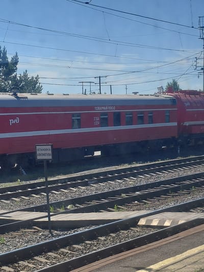 The image shows a red train car with "РЖД" branding on its side, indicating it belongs to Russian Railways. The train car has windows and is positioned on railroad tracks. A signpost with the text "ОСТАНОВКА ЛОКОМОТИВА" (Locomotive Stop) is visible near the train. In the background, there are overhead power lines and trees. The scene appears to be a railway station in the city of Chulym, Russia. The sky is blue with some clouds, suggesting it is daytime. There are no people or specific actions depicted in the image.
