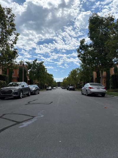 The image depicts a street scene in Irvine, California, under a partly cloudy sky. The perspective is low, close to the asphalt road which shows some dark tar repairs and what appears to be a light-colored spill. On the left side of the road, a dark grey Audi SUV is parked. Further down the street on the left, several other cars are parked, and trees line the sidewalk. The houses on the left are Mediterranean-style, with orange stucco exteriors and red-tiled roofs. On the right side of the road, a silver sedan is parked. Beyond it, more trees and houses are visible. The street is relatively narrow, and appears to be a residential area.