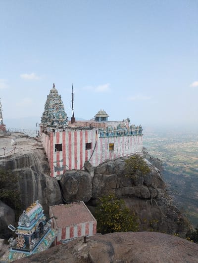 The image displays a Hindu temple complex situated on a prominent rocky outcrop in Kadaladi, India, overlooking a vast, hazy landscape. The primary temple structure, positioned centrally, features walls with vertical red and white stripes and several dark, rectangular window openings. A multi-tiered, ornate gopuram (temple tower) with blue, gold, and white detailing rises from the left side of this building's roof. To its right, the roof is adorned with smaller decorative structures, including a light blue dome. This main structure is integrated into large, grey natural rock formations. Below and to the left, a smaller, intricately decorated shrine with a colorful gopuram-like apex is visible, also built upon the rock. Adjacent to it, a small, cube-shaped building with red and white striped walls rests on a lower rock shelf. Sparse green and yellow flowering vegetation grows around the rock formations. The sky is light blue with scattered white clouds, and the distant background reveals an expansive, flat plain under a light haze, indicative of a significant elevation. No individuals or discernible actions are present.
