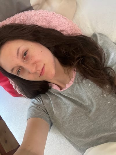 In Popintsi, Bulgaria, a young woman with long, dark brown hair is lying down, looking at the camera. She is wearing a grey t-shirt with small white stars on it and a pink trim around the collar. Her left arm is resting on a white surface, and her head is propped up on a pink fuzzy pillow, which is on top of a grey and white patterned surface. A red pillow is visible behind her head on the left. The environment appears to be indoors, possibly a bedroom, with a white bed or couch as the primary surface. The lighting is soft, suggesting it could be daytime.