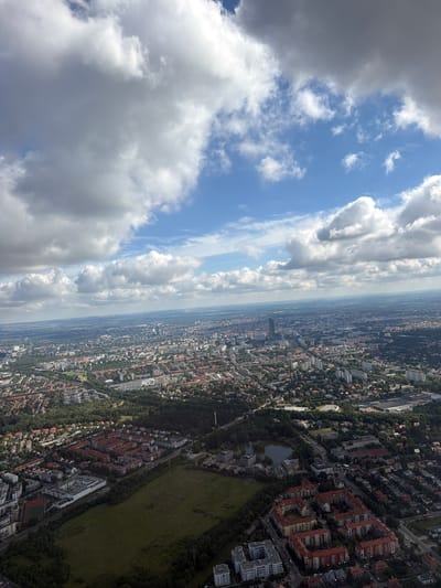 An aerial perspective captures a sprawling cityscape of Raczkowa, Poland, under a partly cloudy sky with prominent cumulus formations. The view encompasses a vast urban expanse with a dense network of buildings, ranging from residential blocks with red-tiled roofs to taller, modern structures in the distance. Numerous trees and green spaces are interspersed throughout the city, including a noticeable park area with a small lake. Infrastructure such as roads and what appears to be a sports field are visible. The image is static and does not depict any people or discernible actions. The overall environment is that of a developed urban area with a mix of residential, commercial, and recreational zones.