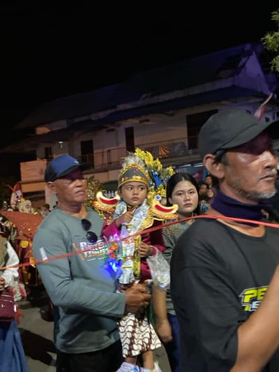 The image depicts a nighttime scene in Kedungbanteng, Indonesia, during a public gathering or parade. In the mid-ground, a man wearing a blue baseball cap and a grey long-sleeve shirt holds a young child. The man's shirt has a graphic design, and black sunglasses are tucked into his collar. The child, prominently featured in the center, is dressed in an elaborate traditional costume, which includes a gold and white feathered headpiece, a red garment with gold trim, and decorative, gold, dragon-like structures on either side, possibly part of a palanquin or support. The child holds a small object with multi-colored LED lights and a transparent plastic bag.

To the immediate right of the child, a woman with dark hair, wearing a greyish-green top, stands with a neutral expression, looking forward. In the foreground, on the right side, a man wearing a black baseball cap, a black t-shirt, and a dark blue neck gaiter is partially visible, facing left. A red rope or string spans horizontally across the mid-ground, passing in front of the man holding the child and behind the foreground man.

The background reveals a multi-story building with a dark blue roof under artificial illumination. An Indonesian flag (red and white) is visible on a pole attached to the building. The overall scene is lit by artificial lights, indicating an evening or night event, with several additional blurred figures suggesting a crowd.
