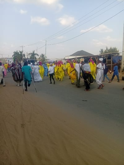 The image depicts an outdoor street scene at Itakpe Junction, Nigeria, under a light blue sky with scattered white clouds. A procession of approximately 20-30 individuals occupies the central and right portions of the frame, moving from left to right.

The ground transitions from an unpaved, sandy surface in the foreground to a paved road further into the midground. The largest group of participants, positioned centrally, consists of approximately ten individuals dressed in vibrant yellow, long-sleeved garments, many featuring elaborate pink or red feather-like head adornments. One individual within this group carries a white, rounded object on their head. To the left of this yellow-clad group, a person wearing a teal-colored top and dark trousers walks with a prosthetic left leg and uses two crutches. Further left, a person in a dark and white striped, possibly ceremonial, costume is partially visible. Several other individuals are dressed in white robes or garments, some with head coverings. On the far right, two children in casual attire are walking.

In the background, utility poles with multiple overhead power lines are visible against the sky. Various buildings line the street, including structures with dark, angular roofs and what appear to be rudimentary market stalls or shelters on the right. Palm trees are present on the left side of the road, and a distant building with a prominent minaret-like feature is visible on the far left. A billboard with green text is partially visible on the left side of the street. The scene is illuminated by natural daylight.