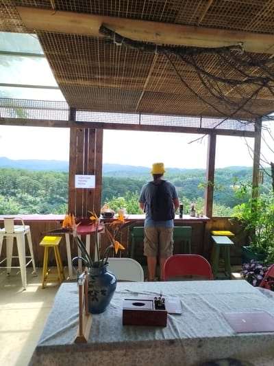 The image depicts an outdoor seating area with a panoramic view of a lush, forested landscape. In the foreground, a table is draped with a patterned tablecloth, and on it sits a blue patterned vase filled with orange and yellow flowers, a wooden napkin holder, and a menu. Behind the table, several chairs are visible, including a red one and two green ones. To the left, a white stool stands near a small yellow stool.
A man wearing a yellow bucket hat, a blue t-shirt, and khaki shorts stands with his back to the camera, looking out at the view. He is wearing a black backpack and holds a silver thermos in his left hand. A sign on a wooden post behind him reads "TIEM KEM TRONG RUNG" in Vietnamese, which translates to "Ice Cream in the Forest." The structure above the seating area is made of wood with a lattice ceiling, and branches hang down. The background features rolling hills covered in dense greenery, indicating a natural or park-like setting, likely in the vicinity of Trường An, Vietnam, known for its scenic beauty. The overall atmosphere is relaxed and natural.