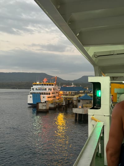 An outdoor, medium-shot, eye-level, vertical, full-color, natural light photograph captures a ferry docked at a pier in the city of Melaya, Indonesia. The ferry, predominantly white with blue accents, is positioned to the left of the frame, its lights reflecting on the dark water. Several piers and structures, including a small blue-roofed building, are visible extending from the shoreline. A mountainous landscape with lush vegetation forms the backdrop under a cloudy, twilight sky. The foreground is occupied by the perspective from another vessel, with a white railing and a green navigation light prominently featured on the right. The scene suggests a port or harbor environment during the evening hours.
