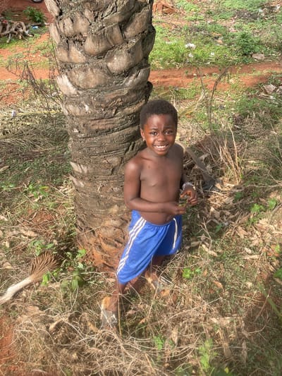 A young, shirtless boy is lying horizontally on the trunk of a felled palm tree in Ogbe, Nigeria. He is smiling and wearing blue shorts with white stripes down the side. The trunk of the palm tree runs vertically and is surrounded by dry grass, small green plants, and scattered leaves. A pair of shoes and a small palm flower are visible in the lower right corner of the frame. The location appears to be an area with natural vegetation and red soil.