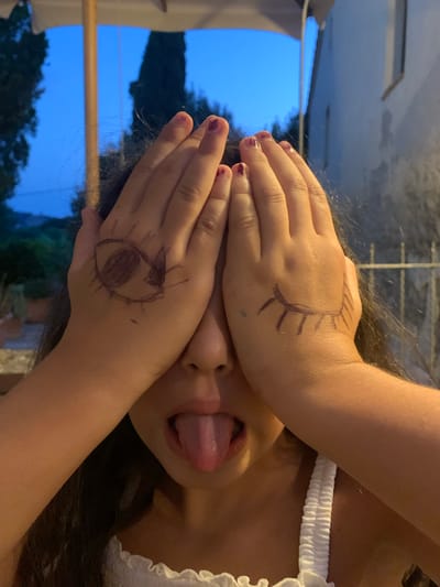 In Bagno Vignoni, Italy, a close-up shot focuses on a young girl with dark hair covering her face, her hands obscuring her eyes, each hand adorned with a drawing of an eye. Her tongue is sticking out from between her lips, indicating playfulness. The background reveals a twilight sky, trees, and a building, suggesting an outdoor setting in the evening.
