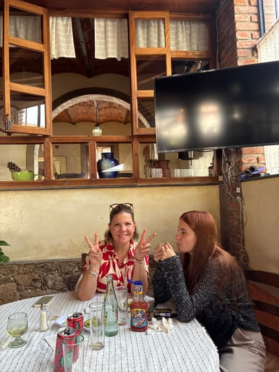 Two women are seated at a table covered with a white and blue striped cloth, laden with bottles, cans, and glasses. The woman closest to the camera is smiling and holding up a peace sign. The other woman is looking down at her hands. They are seated outside, in an outdoor patio with a stone wall. A window with wooden frame panes is next to the table. Through the window, the architectural details are visible suggesting the location is Tepeji de Ocampo, Mexico. Wires are draped along the upper wall, and a television screen is attached.
