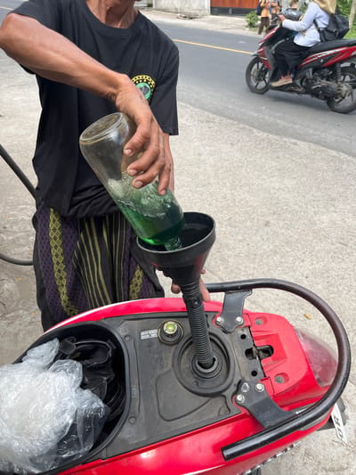 In Tanakawu, Indonesia, an individual is depicted pouring green liquid from a glass bottle into the fuel tank of a red scooter using a black funnel. The individual is wearing a black t-shirt and a sarong with a vertical striped pattern in black, green, and purple. The scooter's storage compartment beneath the seat is visible, containing a plastic bag filled with what appears to be shoes. In the background, a street scene unfolds with a yellow line dividing the road, and two individuals on a red motorcycle are seen riding away. The ground surface is a mix of concrete and asphalt.