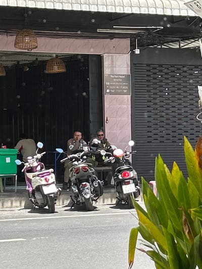 In Surat Thani City Municipality, Thailand, two uniformed individuals, seemingly police officers, are seated on scooters outside a building with a closed metal grate storefront. The officer on the left is wearing a light-colored uniform and appears to be holding something to his ear. The officer on the right wears a similar uniform and has sunglasses on. Two scooters are parked in front of them; one is white and purple, and the other is black and gray. Behind the officers, a sign is visible that reads "The Port Hostel" and lists "Service Fees." A green bin is located to the left of the officers. The building has a tiled facade and a covered walkway with woven light fixtures. Lush green foliage is in the foreground.