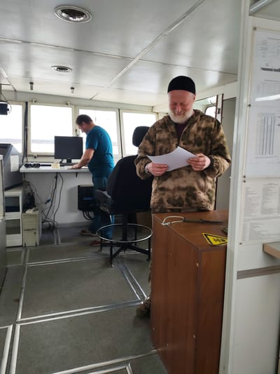 The image depicts the interior of what appears to be a ship's bridge or wheelhouse, characterized by large windows, a functional layout, and specialized equipment. Two men are present, engaged in activities.

On the right, a man with a white beard and a black cap stands, wearing a camouflage-patterned jacket. He is looking down and holding several white papers in his hands, appearing to read or review them. A subtle smile or contented expression is visible on his face. On a wooden cabinet next to him, a sheathed knife with a lanyard and a yellow hazard symbol sticker are visible. A white board mounted on the wall to his right holds several documents with unreadable text.

On the left side of the frame, another man in a blue t-shirt and dark pants is standing at a desk, facing a computer monitor (brand "SAMSUNG") and keyboard. He appears to be operating the computer. On the desk near him are a pair of binoculars and some communication devices, including a walkie-talkie. Below the desk, a white computer tower sits next to various cables, and a black electrical unit labeled "FDP FSZ 105.20" is mounted on the wall. A black office chair with wheels is positioned in front of the desk.

The setting is brightly lit, likely by natural daylight streaming through the numerous windows that offer a view of a distant landscape or water, consistent with a vessel in Rybinsk, Russia. The ceiling is white with visible paneling and circular recessed light fixtures. The floor is grey, textured, and marked with a grid of metal strips; the text "B-1 058" is visible on one of the floor panels under the chair. The overall scene suggests a routine work environment on a marine vessel.