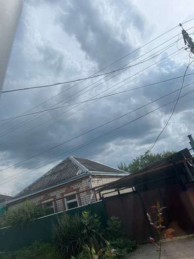 The image shows a cloudy sky over a residential area in Kochubeyevskoye, Russia. In the foreground, a dark brown metal gate is partially visible, with a small rose bush in front of it. Behind the gate, a single-story house with a red brick base and white brick upper walls is visible. The house has a gray, corrugated roof and white-framed windows. A green metal fence runs along the front of the property, separating it from the street. Overhead, numerous power lines crisscross the sky, supported by a utility pole on the right side of the frame. The overall scene depicts a typical residential street on an overcast day.