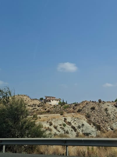 The image depicts a dry, hilly landscape with sparse vegetation under a clear blue sky with a few scattered white clouds. In the mid-ground, a two-story building with a light-colored facade and a tiled roof is situated on a hilltop. The terrain is characterized by rocky slopes and dry grasses. A metal barrier is visible in the foreground, partially obscuring the lower part of the scene. The overall impression is one of a semi-arid environment, likely in a rural or semi-rural area.