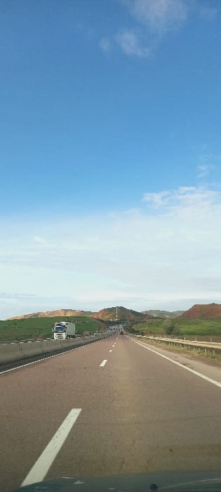 The image displays a daytime highway scene near Majmaa Tolba, Morocco, captured from the interior of a moving vehicle. The central feature is a multi-lane asphalt highway extending into the distance, with visible white dashed lane markings and a solid white line on the right shoulder. The lower portion of the image shows a dark, indeterminate edge of the vehicle's dashboard or windshield.

To the left of the highway, a concrete barrier is visible, beyond which a white semi-truck with a white box trailer is present, traveling in the same direction. To the right of the highway, a metal guardrail runs parallel to the road, with sparse green vegetation immediately beyond it.

In the mid-ground and background, rolling hills characterize the landscape. Hills on the left are predominantly green, while those on the right exhibit a mix of green vegetation and reddish-brown soil. A distinct hill in the central-right distance features a vertical structure, consistent with a communication tower, on its summit. Additional vehicles are discernible on the distant highway lanes, appearing as small forms. The sky occupies the upper portion of the frame, displaying a clear blue with minimal, scattered white cumulus clouds, primarily towards the upper right. The lighting indicates bright daylight conditions. No human figures are explicitly visible.