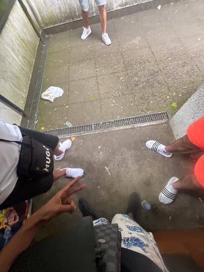The image depicts a group of people standing on a paved surface with a drainage grate in Liestal, Switzerland. There are at least three people visible. One individual wears orange shorts and white Adidas slide sandals with black stripes, with socks. Another person, wearing dark clothing and white Nike shoes with red accents, is making a hand gesture. A third person is partly visible, wearing dark clothing and a white and blue patterned bag. There is a trash bag and loose debris scattered on the ground. The environment appears to be an urban outdoor area.