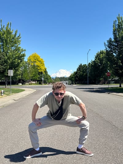 A man with curly brown hair and sunglasses crouches in the middle of a residential street in South Hill, Washington, United States of America. He wears a light green t-shirt, light gray pants, and brown shoes with white soles. A black fanny pack is slung across his chest. His right hand rests on his right knee, and his left arm is bent at the elbow. The street is paved and lined with trees on both sides. In the background, a snow-capped mountain, likely Mount Rainier, is visible under a clear blue sky. A stop sign is posted on the right side of the street, and a fire hydrant is on the left. The sun casts long shadows on the asphalt.