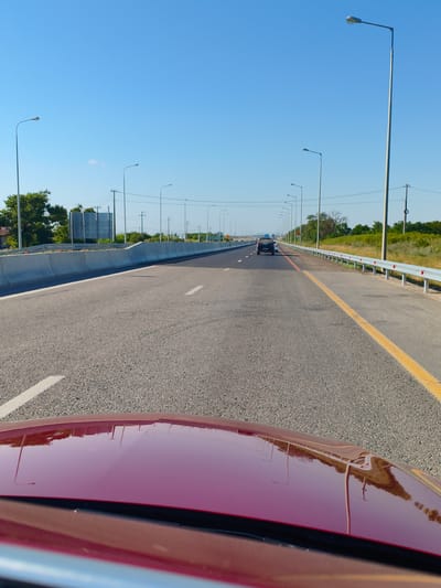 A first-person perspective shot captures a view from inside a car on a clear, sunny day, traveling on a highway. The highway is paved and marked with white and yellow lines, with a concrete barrier on the left and a guardrail on the right. Numerous streetlights line the sides of the road, and vegetation is visible beyond the barriers. In the distance, two cars are on the highway, one in each lane, with the rear of the closest vehicle visible. The foreground shows the reflection of the sky and streetlights on the polished hood of the car. The location is identified as Piatikhatki, Russia.