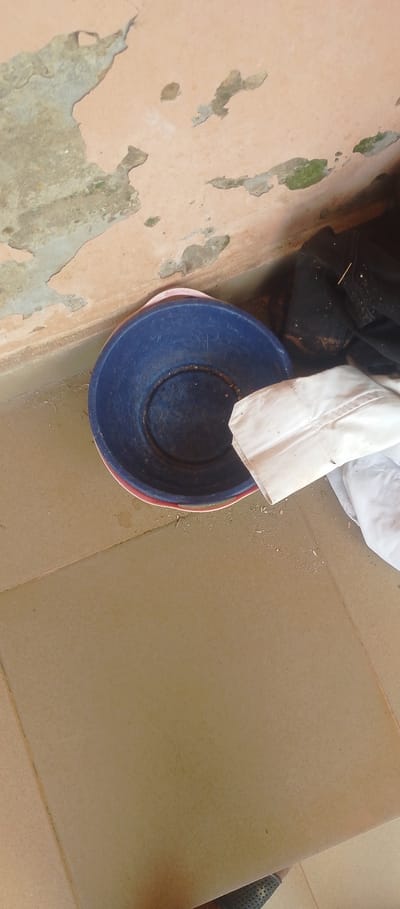 A medium shot captures a blue plastic basin with a red rim resting on a tiled floor in front of a peeling, light-colored wall in Achara Ndiagu Omegio, Nigeria. A crumpled white cloth lies partially over the basin, and dark fabric, possibly clothing, is visible behind it. The wall exhibits signs of wear with patches of green discoloration and exposed concrete. The floor tiles are a light brown, and small debris is scattered on the floor and in the basin. The scene appears to be indoors or under a covered outdoor space.