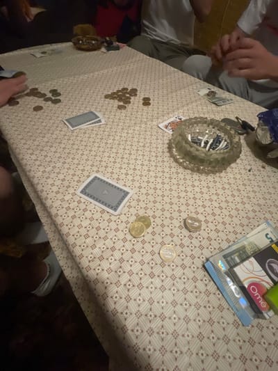 The image shows a table with a patterned tablecloth. Several people are sitting around the table, visible only partially. There are coins scattered on the table, along with playing cards, cash, condoms, a lighter, and an ashtray filled with cigarette butts. There is no clear indication of the location in Krumovgrad, Bulgaria.