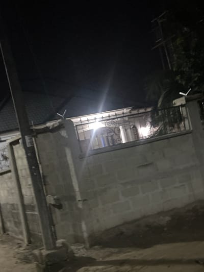 An eye-level, rotated view of a building at night, illuminated by a bright light inside. The building is constructed of light-colored bricks or concrete blocks, with a metal railing visible on an upper level. Foliage is visible at the top left, contrasting with the darkness of the sky. The building's structure includes horizontal beams and possibly a lower level partially visible in the lower part of the frame. The overall image suggests an outdoor scene at dusk or night, with a combination of structural details and natural elements.