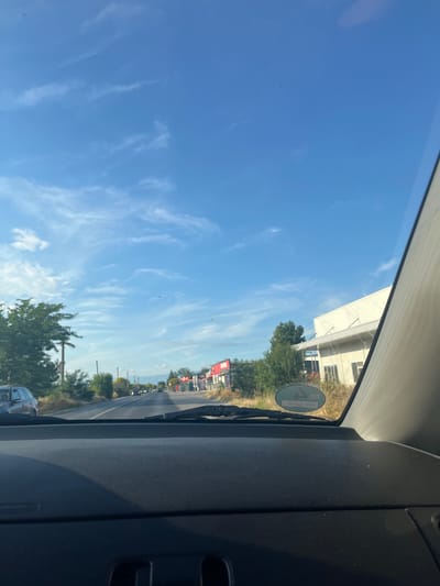 The image captures a view from inside a car driving down a road in Graf Ignatievo, Bulgaria. The sky is a bright blue with scattered wispy white clouds. On the right side of the road, there are several buildings with white exteriors, some appearing to be commercial establishments. Trees and vegetation line the sides of the road. In the distance, the road stretches out, with a few cars visible, and mountains can be seen on the horizon. The dashboard of the car is visible in the foreground.