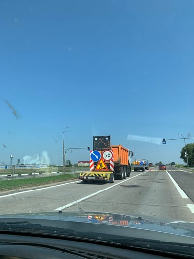 The image captures a scene on a highway in Азерскі сельскі Савет, Belarus, on a clear day. In the foreground, the dashboard of a vehicle is visible, indicating the perspective of a driver. The road ahead features multiple lanes, with a red car in the distance and other vehicles, including a large orange dump truck with a trailer carrying road construction signs, further ahead. The dump truck displays several traffic signs, including a speed limit of 50 and a sign indicating road work. Traffic lights are visible on poles alongside the road, and construction cones are present, suggesting ongoing road maintenance or construction. The surrounding environment includes grassy verges, trees, and buildings in the distance. The sky is a clear blue, with some light smudges on the windshield.