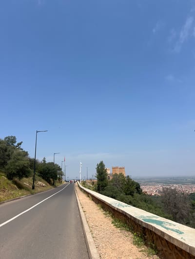 The image shows a paved road winding along a hillside in Beni Mellal, Morocco. A white line divides the road. To the right, a rough stone wall borders the road with vegetation. Beyond the wall is a view of the city. To the left of the road are trees and streetlights. A distant building with a tower is visible. The sky is blue.