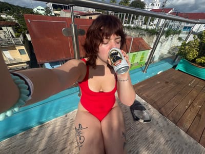A woman in a red swimsuit poses upside down, holding a beer can to her forehead. She stands on a structure with a blue railing, with buildings visible in the background. The woman has dark hair and tattoos on her legs. Her hand is visible in the foreground wearing a bracelet. To the left, there's a wooden surface with a sneaker. The camera angle is unusual, emphasizing the upside-down perspective.