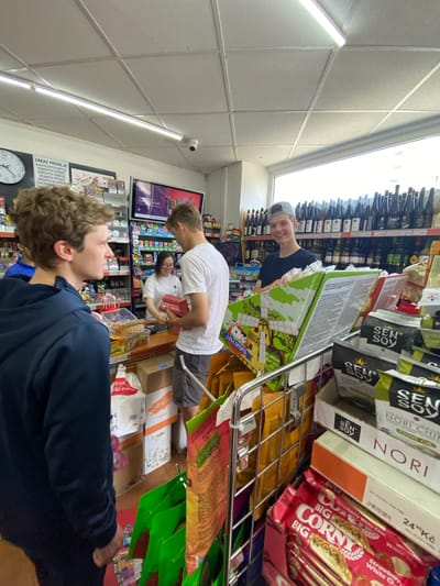 The image shows three young men in a convenience store in Hradec Králové, Czechia. The first man, wearing a navy blue hoodie, is in the foreground, facing away from the camera. In the middle ground, a second man wearing a white t-shirt is holding red items at the checkout counter where an Asian woman in a white shirt is working. In the background, a third man wearing a dark t-shirt and baseball cap is looking at the camera. The store shelves display a variety of products, including wine bottles, snacks such as Sen Soy Nori chips and Corny Big bars, and other packaged goods. A sign on the wall reads "ZÁKAZ PRODEJE ALKOHOLICKÝCH NÁPOJŮ" (Prohibition of the sale of alcoholic beverages), along with other restrictions. A clock and an advertisement for Glo are also visible on the wall.