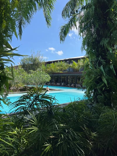 A scene in Petulu, Indonesia shows a partially obscured view of a swimming pool and a multi-story building, framed by dense, lush vegetation. The pool's turquoise water contrasts with the surrounding greenery. A person stands near the edge of the pool. The building features a series of balconies or terraces with visible furniture, suggesting a resort or residential complex. The sky above is blue with scattered clouds. Tropical plants dominate the foreground, creating a natural border around the view.