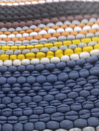 This is a close-up, slightly blurred photograph of a textured woven fabric, likely a rug or textile. The fabric is composed of horizontal rows of rounded, bulbous stitches. The colors present are dark grey, brown, light grey, orange, yellow, white, and a prominent dark blue. The blue section occupies the majority of the lower half of the image, with rows of densely packed blue stitches. Above this are alternating rows of white, yellow, orange, and grey stitches. Further up, brown and dark grey rows are visible. The texture suggests a tactile, woven material. There are no people, animals, or any discernible environmental context suggesting a specific location. The image provides no information related to the city of Balâ, Turkey.