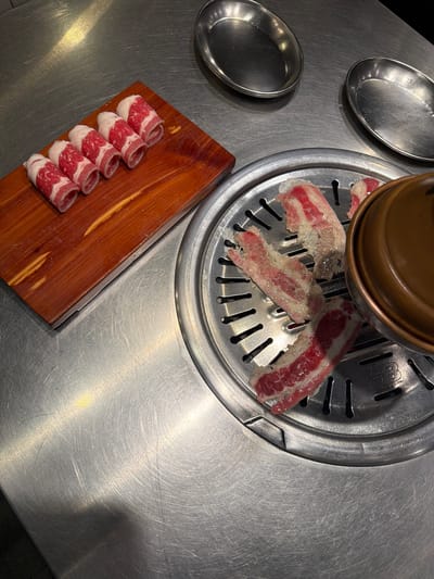The image depicts a close-up, high-angle shot of a Korean barbecue setup. In the foreground, raw, thinly sliced beef rolls are arranged on a wooden board. To the right, more slices of raw beef are placed on a metal grill plate, which is part of a circular barbecue unit. A copper-colored lid is partially visible to the right of the grill. In the background, two oval-shaped metal dishes sit on a stainless steel surface. The overall environment suggests a restaurant or a home cooking setup for Korean barbecue, common in urban centers like Cebu City, Philippines, where such dining experiences are popular. No people are visible in the frame. The lighting is bright, highlighting the details of the raw meat and the metallic surfaces.