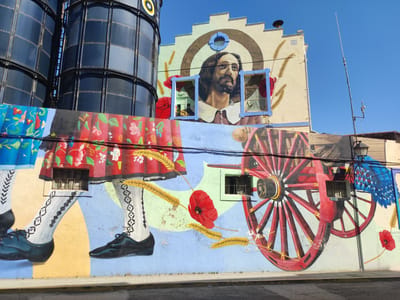This media file captures a vibrant mural painted on a building facade in Valencia de Alcántara, Spain. The mural depicts a collection of folk-art inspired elements. Dominating the upper portion of the mural is a large portrait of a man with a beard, framed by a halo-like arch and surrounded by depictions of wheat stalks and red poppies. Two blue-framed windows interrupt the portrait, adding a surreal quality. Below this, the mural continues with stylized imagery of traditional folk attire, including colorful patterned skirts, patterned white stockings with black diamond designs, and black lace-up shoes. A large, red wagon wheel is a prominent feature on the right side of the mural, with a second, partially obscured wheel visible behind it. Other elements include depictions of blue feathers and more red poppies and wheat stalks. Two small, barred windows are integrated into the mural, appearing as if they are part of the painted scene. In the background, two large, dark, cylindrical silos rise vertically. The scene is illuminated by bright daylight under a clear blue sky, suggesting a sunny day. The overall impression is one of cultural celebration and artistic expression. Visible text includes "THEO" and "ACUA" written in script on the building facade above the main mural.