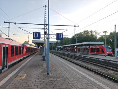 The image depicts a train station platform in Memmingen, Germany, at what appears to be late afternoon or early evening, given the lighting. Two red regional trains are visible, one parked on the left platform and another on the right platform, with an empty track between them. The train on the left is a multi-carriage unit with visible doors and windows. The train on the right is a locomotive-hauled train, also red, with its front end facing the camera.
The platforms are paved with light-colored rectangular bricks, and a white line delineates the edge of the platform. Overhead power lines for the trains are prominently visible, supported by metal poles. Blue signs with the letter "D" are mounted on poles, indicating platforms or tracks. A covered waiting area with seating is present on the right platform. A trash bin is also visible on the left platform.
In the background, trees line the area beyond the tracks, and some buildings and infrastructure associated with a train station, such as signals and more power line supports, are scattered throughout. A few individuals are casually present on the platform, one standing near the train on the right and others potentially inside the train carriages or further down the platform, though they are not the main focus. The sky is clear with some wispy clouds. The scene is orderly and functional, typical of a German public transportation hub.