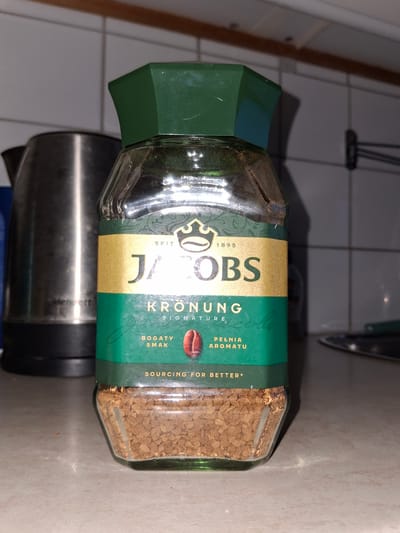 A jar of Jacobs Kronung instant coffee is placed on a countertop, likely in a kitchen setting in Åsgårdstrand, Norway. The jar is predominantly green and gold, with the brand name "JACOBS" prominently displayed in gold lettering. The coffee inside is visible at the bottom of the glass jar, appearing as small brown granules. To the left of the coffee jar, a silver electric kettle is partially visible. The background consists of tiled walls and a wooden surface, suggesting a domestic environment. The lighting in the scene is somewhat dim, with shadows cast on the countertop.