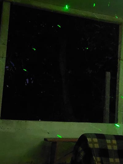 The image depicts a night scene viewed through what appears to be a window or an opening, possibly from an indoor perspective looking outwards. The dominant feature is the darkness, with several small, bright green lights scattered throughout the frame. These lights are irregular in shape, some appearing as streaks or dashes, while others are more like small dots or arcs. The background is very dark, with indistinct shapes that suggest trees or foliage. The foreground is dimly lit with a greenish hue, showing a textured surface that could be a wall or a balcony railing. On the right side, there are vertical elements that resemble wooden posts or framing. In the lower right quadrant, a portion of a plaid blanket draped over an unseen object is visible. The lighting suggests artificial illumination, possibly from a projector or decorative lights. No people or specific actions are discernible. The location is identified as Areni, Armenia, suggesting this is a view from within or near a structure in that region at night.