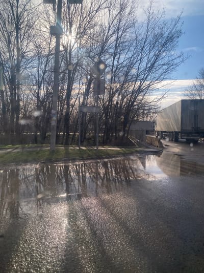 The image depicts an outdoor scene in Oktyabrskiy, Russia, on what appears to be a clear day with low sunlight. The foreground features a substantial puddle covering a dark asphalt surface, reflecting the bare trees and bright sky. Sun glare is prominent on the puddle's surface. To the left, the asphalt transitions to a muddy, grassy shoulder. A tall utility pole stands slightly left of center, equipped with several mounted boxes, and is silhouetted against the bright sun, which causes significant lens flare. Behind the pole and extending across the midground is a dense line of leafless deciduous trees. To the right of the utility pole, another smaller post displays a dark, rectangular sign pointing left, with an additional sign above it. Further right, a small, light-colored structure is partially visible behind the trees. A concrete or wooden ramp extends from the left edge of the road, meeting the puddle area. In the right background, a light-colored semi-trailer truck is parked or moving slowly, with sunlight reflecting off its trailer. Another truck is partially visible behind it. The sky in the upper right quadrant is blue with scattered white clouds. No people or animals are present.