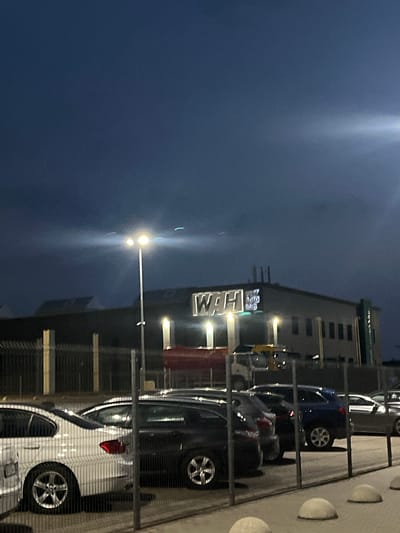 The image shows a dark, possibly cloudy evening scene in Strumivka, Ukraine, with artificial lighting illuminating a parking lot and a building. A prominent illuminated sign on the building reads "WAH AUTO HUB". Several vehicles are parked in the lot, including a white sedan in the foreground. A tall street light with two bright bulbs is visible on the left side of the frame, casting light onto the scene. A metal fence with vertical posts and horizontal bars separates the viewer from the parking lot and vehicles. The building appears to be a commercial establishment. The sky is dark blue, suggesting it is either dusk or night.