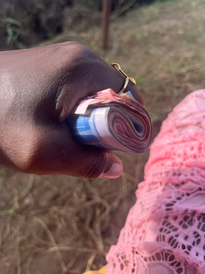 The image presents a close-up, high-angle view of a person's hand holding a substantial roll of banknotes, set outdoors in Aghalokpe, Nigeria.

The main subject is a person's hand, characterized by its dark skin tone. A distinctive gold-colored ring, featuring a cross-like design, is worn on one of the fingers. The hand is firmly grasping a thick bundle of currency, rolled tightly into a cylindrical shape. While specific denominations or text on the banknotes are not discernible due to their rolled state and partial blur, colors such as blue, red, and purple are visible on the edges.

In the upper right background, a soft pink, lacy fabric with an intricate, open-work pattern is partially visible. This fabric appears to be part of clothing or an accessory belonging to another individual or item in the scene. The rest of the background is heavily out of focus, depicting muted tones of green and brown, suggesting an outdoor environment with natural elements like grass or dirt.

The scene is illuminated by bright daylight, casting a discernible shadow across the holding hand, indicating sunny weather. The act of holding such a roll of money implies a potential transaction, a display of wealth, or preparation for expenditure. No specific text is visible on the currency, the ring, or the background elements.