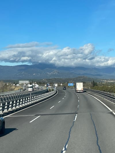 The image displays a multi-lane highway scene in the vicinity of Pont de Molins, Spain, under a bright blue sky with scattered cumulus clouds partially obscuring distant, vegetated mountains. The perspective is from a vehicle traveling forward on the highway. The asphalt roadway features white dashed and solid lane markings, with visible longitudinal cracks in the immediate foreground. A silver metal guardrail system lines the left side of the road. Multiple vehicles, primarily white tractor-trailer trucks and several passenger cars, are observed traveling in both directions. One white truck is prominent in the right-hand lane, moving away from the viewer. In the mid-distance, a blue overhead road sign with white text, indicating "LE JONQUERA," is visible. The surrounding environment consists of sparse roadside vegetation and rolling hills leading to the mountain range. No individual people are discernible.