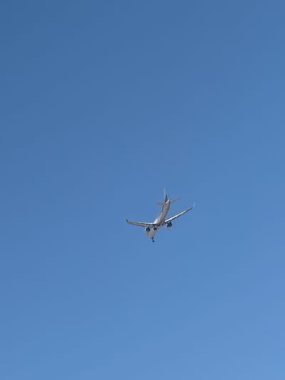 An airplane is captured in mid-flight against a clear blue sky, its undercarriage visible as it appears to be descending or banking. The airplane is predominantly white with colored accents on its tail, featuring green, white, and red stripes. The image was taken in the vicinity of Slanchevo, Bulgaria, suggesting the aircraft might be approaching or departing from a nearby airport. The vast expanse of the sky dominates the frame, with no other discernible elements in the environment.