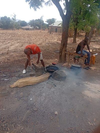 Two men are standing in a dirt lot in Assai Sabongida, Nigeria, at coordinates (latitude: 8.864, longitude: 8.442). There are several trees in the background, including a large tree on the left side of the image and a smaller tree on the right side of the image. There is a small building in the background, which is likely a house. The man on the left is wearing a red shirt and white shorts. He is bending down, looking at something on the ground. He is holding a piece of wood in his right hand. The man on the right is wearing a blue shirt and brown pants. He is sitting on a bench that has a small fire in front of it. He is holding a piece of wood in his right hand. There is a large rock in the center of the image. The men are in an outdoor setting, and it looks like they are cooking. The man on the right appears to be watching the fire. There is smoke coming from the fire. The ground is covered in dirt and small rocks. There are several other objects in the scene, including a large log, a small pot, and a small bucket. The scene is illuminated by natural light.
