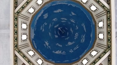 This image is a low-angle, full shot of a domed ceiling decorated with a blue sky and white clouds. The dome is octagonal and surrounded by an ornate border featuring green and gold geometric patterns. Along the perimeter of the dome are eight evenly spaced windows, each with a decorative frame and emitting a bright, white light. In the center of the dome, a white, slender object, possibly a light fixture or architectural element, hangs down from the apex. The overall impression is of an interior space with an artistic, sky-like ceiling. The location is the city of Beunyot, Indonesia.
