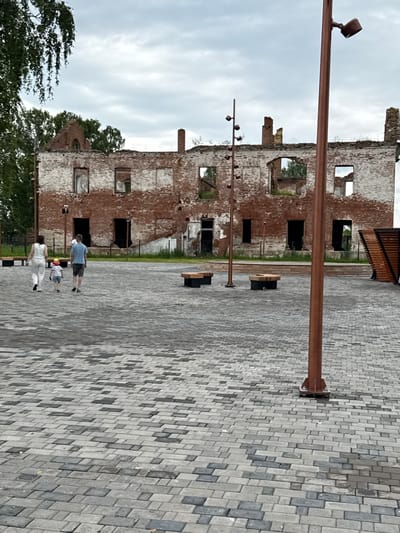 The image shows a family walking in a public square with a ruined building in the background. A couple, a man and a woman, are walking with their young child in the center of the frame. The child is wearing a yellow hat. They are walking away from the camera on a paved walkway. In the background, the remains of a brick building are visible, with broken windows and crumbling walls. The sky is overcast. The location is identified as the city of Verkhotor, Russia.